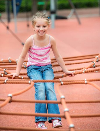 Téléchargez les photos : Heureuse belle petite fille souriante jouant au terrain de jeu et grimpant les cordes en plein air - en image libre de droit