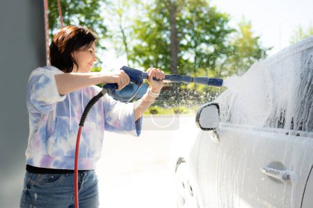 Téléchargez les photos : Fille laver une voiture dans une station de lavage de voiture libre-service avec de la mousse de lavage. Laver la station libre-service de voiture. - en image libre de droit