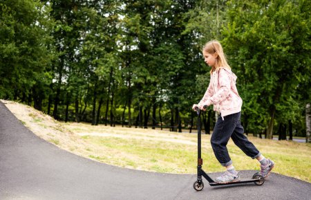 Téléchargez les photos : Preteen fille équitation scooter dans le parc. Enfant féminin mignon avec véhicule écologique à l'extérieur - en image libre de droit
