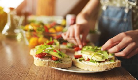 Photo for Girl hands preparing sandwiches with avocado, tomatoes and greens at kitchen closeup. Vegetarian bruschettas with fresh vegetables and arugula - Royalty Free Image