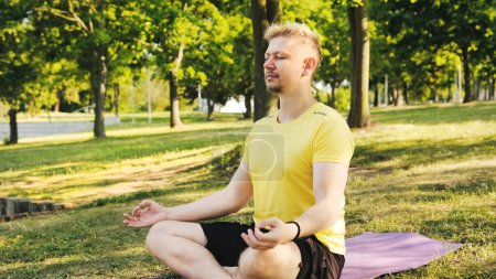 Téléchargez les photos : Jeune homme assis sur un tapis de yoga en position Lotus se préparant à pratiquer le yoga et la méditation à l'extérieur. Fitness et concept sain. - en image libre de droit