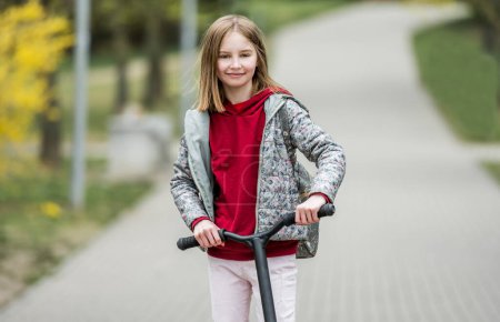 Téléchargez les photos : Petite fille mignonne chevauchant un scooter sur un chemin dans le parc, souriant et regardant dans la caméra - en image libre de droit