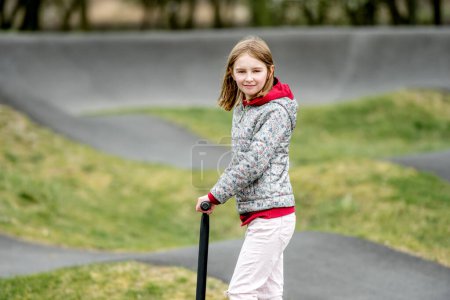 Téléchargez les photos : Belle petite fille monte un scooter dans un parc de balade extrême, sentant et regardant dans la caméra - en image libre de droit