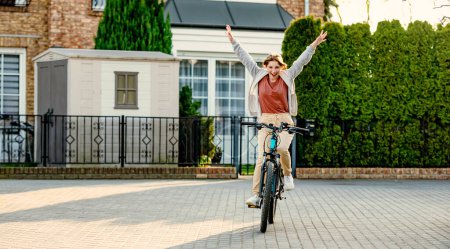 Photo for Funny girl riding a bike in the evening with rising hands up. Young girl joking and having fun. Eco-friendly urban transport. - Royalty Free Image