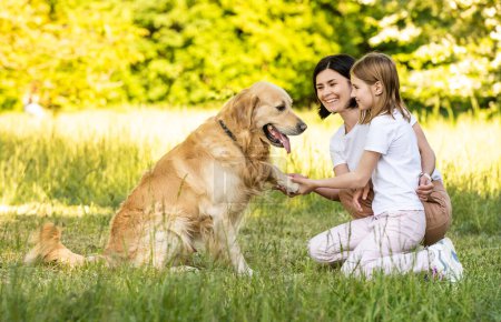 Foto de Madre y linda hija jugando con adorable perro golden retriever al aire libre - Imagen libre de derechos