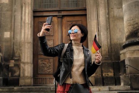 Photo for Attractive Female Tourist Walks With Germany Flag In Historical Center Of Dresden, Taking A Selfie - Royalty Free Image