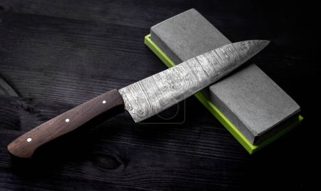 Téléchargez les photos : Grand couteau de cuisine repose sur l'affûtage de la pierre sur la table de cuisine sur fond noir - en image libre de droit