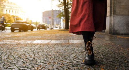 Photo for Female In Long Red Skirt And Black Boots Walks Along Autumn City Street After Rain - Royalty Free Image
