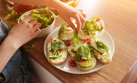 Téléchargez les photos : Fille prépare des sandwichs avec avocat, légumes et légumes verts à la cuisine. Brosses végétariennes aux légumes frais et roquette - en image libre de droit