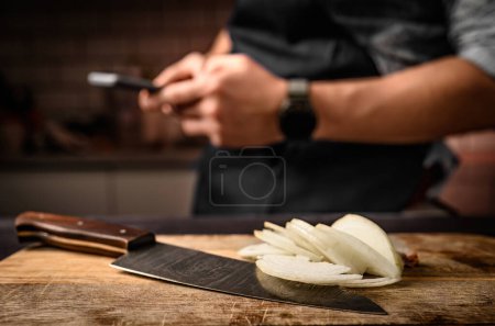 Téléchargez les photos : Chef mains tenant smartphone à la cuisine et oignon tranché sur planche de bois. Homme pendant la pause de cuisson avec téléphone portable et légumes amers - en image libre de droit