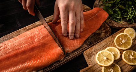 Téléchargez les photos : Les mains principales coupent le filet de saumon avec le couteau sur la table en bois à la cuisine. Homme cuisson oméga rouge poisson pour un dîner sain avec des citrons - en image libre de droit