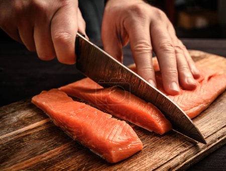 Photo for Chief cutting a salmon fillet into pieces for cooking with a big kitchen knife for healthy dinner - Royalty Free Image