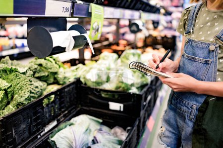 Foto de Niña bonita comprando verduras con lista de compras en el supermercado y revisando productos con notas. Hermosa niña preadolescente femenina en la tienda de comestibles - Imagen libre de derechos