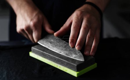 Foto de Hombre afilando cuchillo de cocina con una piedra de afilar se destaca contra fondo negro - Imagen libre de derechos