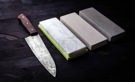 Photo for Big Kitchen Knife Rests On Kitchen Table, Accompanied By Set Of Sharpening Whetstones, All Against Black Background - Royalty Free Image