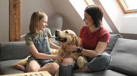 Photo for Pretty girl and preteen child family petting golden retriever dog sitting on sofa. Mother and daughter with purebred pet doggy at home smiling - Royalty Free Image