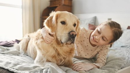 Photo for Cute kid girl petting golden retriever dog lying in bed and smilling in morning. Beautiful preteen kid with purebred pet doggy labrador wake up at home - Royalty Free Image