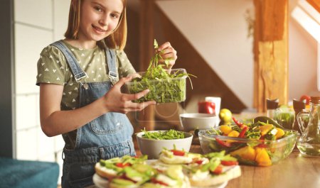 Photo for Girl child holding tray with green arugula at kitchen and smiling. Pretty female kid with vegetables and plants preparing vegetarian lunch - Royalty Free Image
