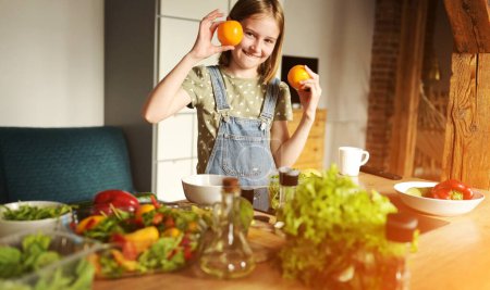 Foto de Niña sosteniendo naranjas en la cocina y sonriendo. Niño bastante hembra con cítricos y verduras preparando almuerzo vegetariano - Imagen libre de derechos