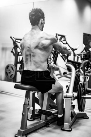 Téléchargez les photos : Forte musclé bodybuilder homme faisant retour avec la machine rangée assis dans une salle de gym moderne - en image libre de droit