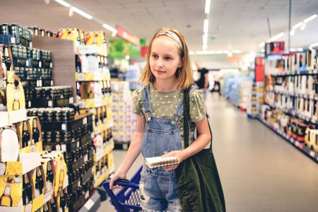 Téléchargez les photos : Jolie fille enfant achetant avec liste de courses au supermarché. Belle fille préadolescente dans le département des boissons alcoolisées avec une diversité de boissons - en image libre de droit