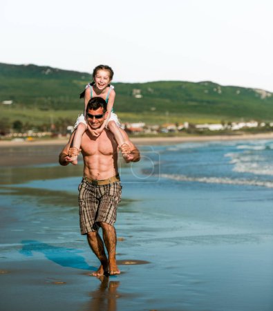 Téléchargez les photos : Père tenant sa fille sur les épaules au bord de la mer et courant. Père de famille et joli enfant souriant à la plage en été - en image libre de droit