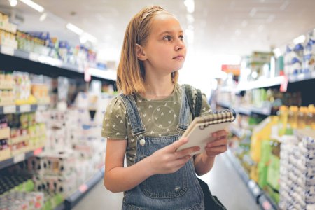 Foto de Niña bonita comprando con lista de compras en el supermercado y mirando estante de producción. Hermosa niña preadolescente femenina en la tienda de comestibles - Imagen libre de derechos