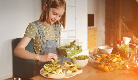 Téléchargez les photos : Fille préparer des sandwichs avec avocat, tomates cerise et roquette à la cuisine. Brosses végétariennes et salade aux légumes frais et légumes verts - en image libre de droit