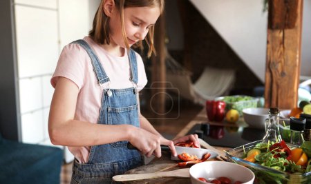 Foto de Linda niña cocinando una ensalada de verduras en la cocina, cortando pimentón rojo en una tabla de cortar - Imagen libre de derechos
