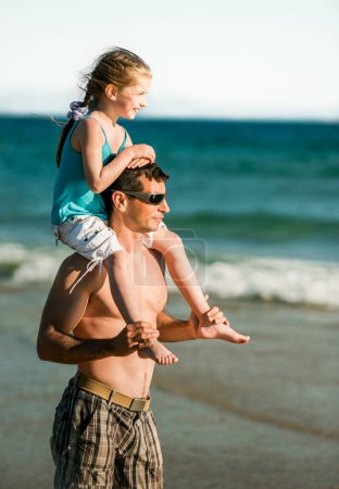 Foto de Padre sosteniendo a su hija sobre hombros en la costa del mar y mirando a la distancia. Padre de familia y niño pequeño y bonito en la playa en verano - Imagen libre de derechos