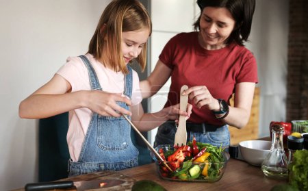 Foto de Hermosa niña con su madre cocinando una ensalada de verduras frescas saludables en la cocina, madre enseña a su hija a cocinar un alimento saludable - Imagen libre de derechos