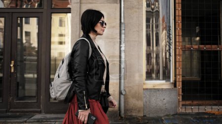Foto de Mujer atractiva en gafas de sol camina por la calle de la ciudad con la mochila, irradiando confianza y estilo - Imagen libre de derechos