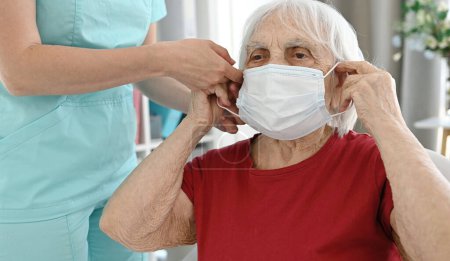 Foto de Enfermera ayuda a la mujer mayor a ponerse una máscara de protección médica durante el período de enfermedades virales y respiratorias - Imagen libre de derechos