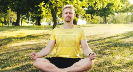 Foto de Joven sentado en una esterilla de yoga en posición de Loto con los ojos cerrados preparándose para practicar yoga al aire libre. Fitness y concepto saludable. - Imagen libre de derechos