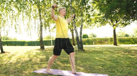 Téléchargez les photos : Jeune homme athlétique faisant des exercices pour les épaules avec la bande élastique sur une herbe À l'extérieur, activités sportives saines et concept de remise en forme - en image libre de droit