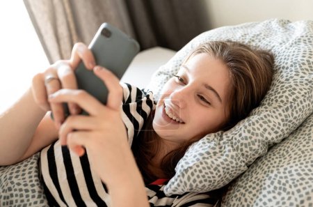 Foto de Adolescente chequeando redes sociales y mensajes en un teléfono inteligente acostado en una cama - Imagen libre de derechos