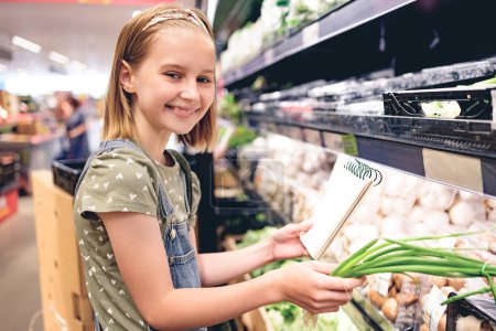 Foto de Niña bonita comprando cebolla verde con lista de compras en el supermercado. Hermosa niña preadolescente femenina eligiendo verduras en la tienda de comestibles - Imagen libre de derechos