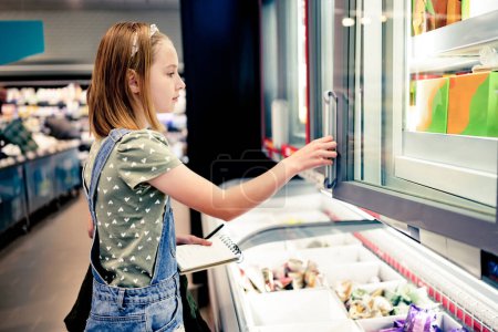 Téléchargez les photos : Jolie fille enfant achetant avec la liste d'achats dans le supermarché et les produits à la recherche dans le réfrigérateur. Belle fille préadolescente dans l'épicerie - en image libre de droit