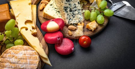 Foto de Diferentes tipos de queso servido con uva y tomate para una nutrición gourmet. Set de brie ecológico y camembert con delicatessen frutos secos sobre mesa negra - Imagen libre de derechos