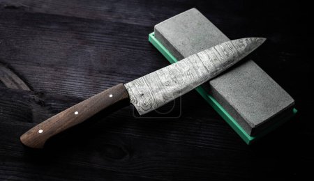 Photo for Sharp Chef's Kitchen Knife And Sharpening Stone Lie On Dark Table - Royalty Free Image