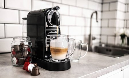 Foto de Máquina de café con cápsulas preparando capuchino en taza transparente en la cocina doméstica en casa. Cafetera expreso cafeína y bebida italiana cremosa - Imagen libre de derechos