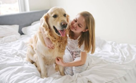 Foto de Dulce niña abrazando un encantador perro Golden Retriever en una cama en la mañana - Imagen libre de derechos