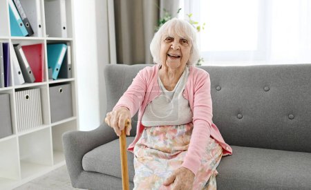 Foto de Mujer mayor sentada en un sofá de la sala de estar con bastón riendo y mirando a la cámara - Imagen libre de derechos