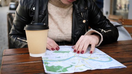 Foto de Elegante turista femenina comprueba la ruta turística de la ciudad en el mapa mientras toma café en el café de la calle - Imagen libre de derechos