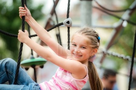 Téléchargez les photos : Portrait de mignonne petite fille souriante jouant sur le terrain de jeu et grimper les cordes en plein air - en image libre de droit