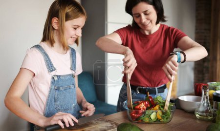 Foto de Hermosa niña con su madre cocinando una ensalada de verduras frescas saludables en la cocina - Imagen libre de derechos