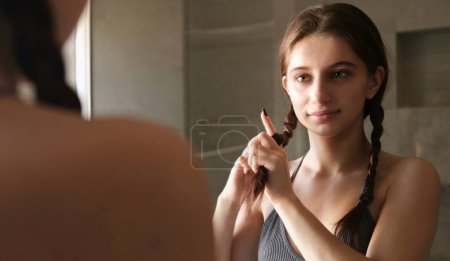 Téléchargez les photos : Adolescente brossant et tressant ses cheveux dans le miroir dans le cadre de sa routine du matin - en image libre de droit