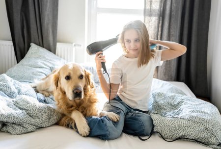 Foto de Feliz niña secando el cabello con un secador de pelo, sentado con el perro en una cama - Imagen libre de derechos