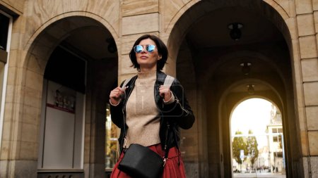 Foto de Atractiva mujer con gafas de sol camina en la ciudad con mochila, turista femenina mirando a su alrededor - Imagen libre de derechos