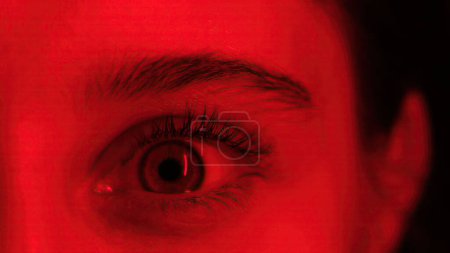 Photo for Young Girl Widely Opens Her Big Eye In Red Light, Seen In A Close-Up View - Royalty Free Image